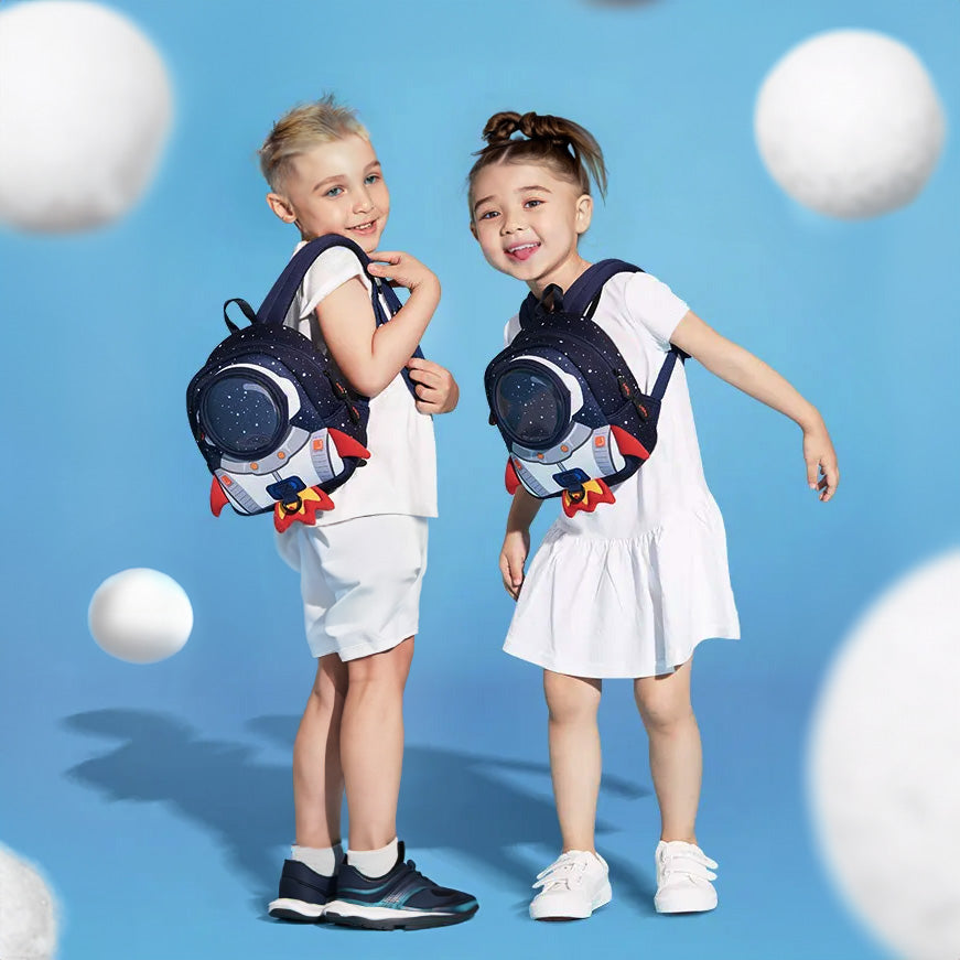 Two children, a boy and a girl, wearing white outfits, are each carrying a blue backpack with a spaceman design. They are standing against a blue background with white orbs around them.